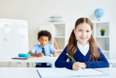 Drawing by desk Adorable schoolgirl with crayon sitting by desk with classmate on background
