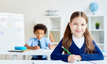 Adorable schoolgirl with crayon sitting by desk with classmate on background